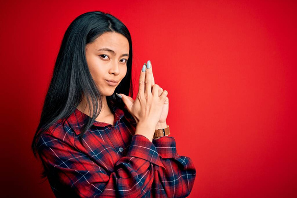 Fiatal gyönyörű kínai nő visel alkalmi ing felett elszigetelt piros háttér Fogása szimbolikus fegyver kézzel gesztus, játék gyilkos fegyverek, dühös arc - Fotó, kép
