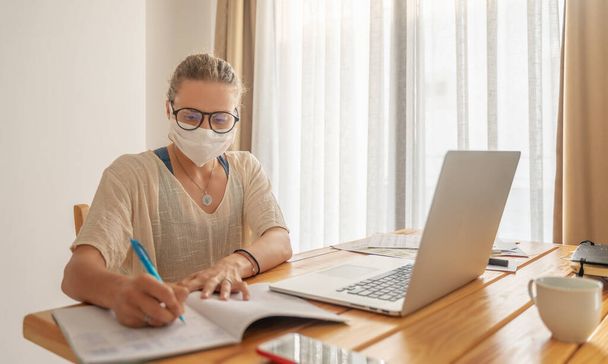 Serious young woman in a medical mask working at a laptop, sits at a table and makes notes in a notebook. Health Safe Work and Education - 写真・画像