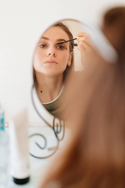 όμορφο κορίτσι κάνει make-up και κοιτάζει στον καθρέφτη. - Φωτογραφία, εικόνα