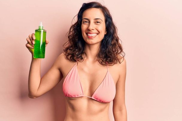 Young beautiful hispanic woman wearing bikini holding aloe vera lotion looking positive and happy standing and smiling with a confident smile showing teeth  - Photo, Image