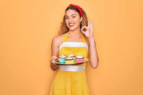 Красива пін-ап жінка з блакитними очима тримає шоколадні кекси на жовтому тлі, роблячи знак з пальцями, відмінний символ
 - Фото, зображення