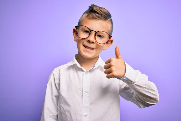 Ein kleines kaukasisches Kind mit blauen Augen, Brille und weißem Hemd vor violettem Hintergrund und glücklichen Daumendrücken mit der Hand. Zustimmender Blick in die Kamera zeigt Erfolg. - Foto, Bild