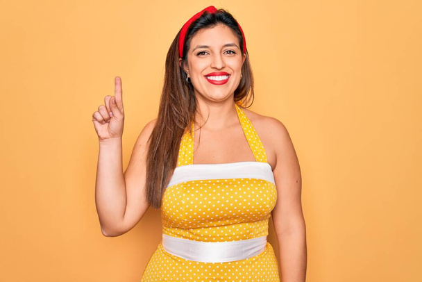 Junge hispanische Pin-up-Frau trägt Mode sexy 50er Jahre Stil auf gelbem Hintergrund zeigt und zeigt mit Finger Nummer eins, während sie selbstbewusst und glücklich lächelt. - Foto, Bild