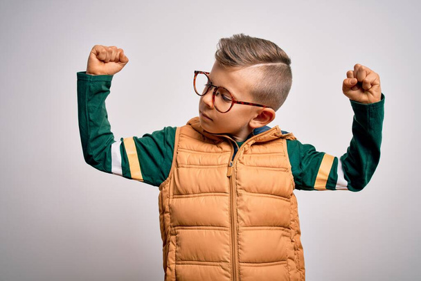Nuori valkoihoinen poika sinisilmäinen yllään talvitakki ja älylasit osoittaa käsivarsien lihaksia hymyilevä ylpeä. Toimivuuskonsepti. - Valokuva, kuva