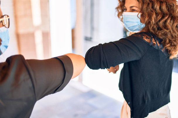 2人の美しい友人がコロナウイルスの予防に新しい肘挨拶をしています。町並みに医療用マスクを着用. - 写真・画像