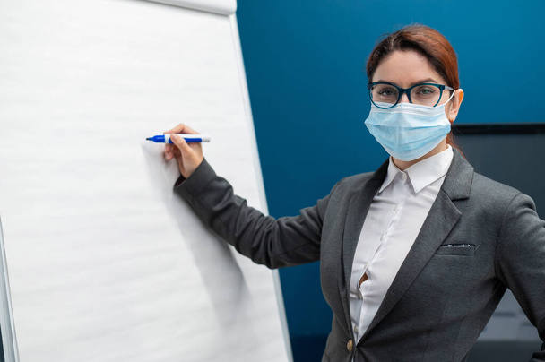 A woman in a medical mask leads a seminar in a conference hall. Business coach writes on a paper white board in the office. Female employee demonstrates presentation while maintaining social distance. - Фото, изображение