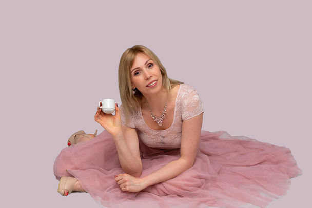 Schöne Frau mit Tasse Tee oder Kaffee. Studioporträt einer verträumten blonden Frau in einem rosafarbenen luftigen Rock, die eine Tasse in der Hand hält und frisches leckeres Getränk trinkt - Foto, Bild