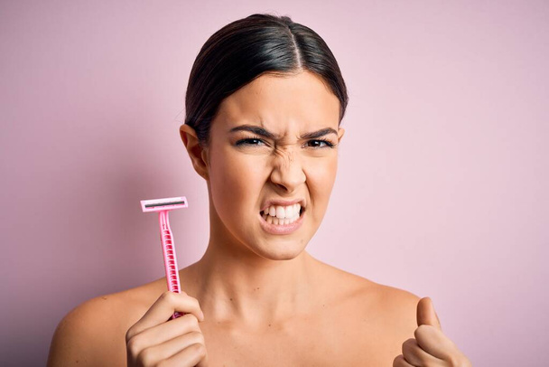 Young beautiful girl using shaver for depilation standing over isolated pink background annoyed and frustrated shouting with anger, crazy and yelling with raised hand, anger concept - 写真・画像