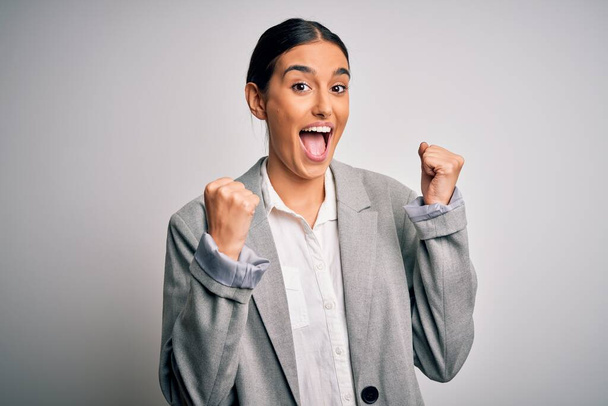 Young beautiful brunette businesswoman wearing jacket standing over white background celebrating surprised and amazed for success with arms raised and open eyes. Winner concept. - Photo, Image