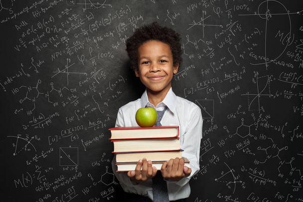Smart black child student smiling and holding books and green apple against blackboard background with science and maths formulas - Photo, Image