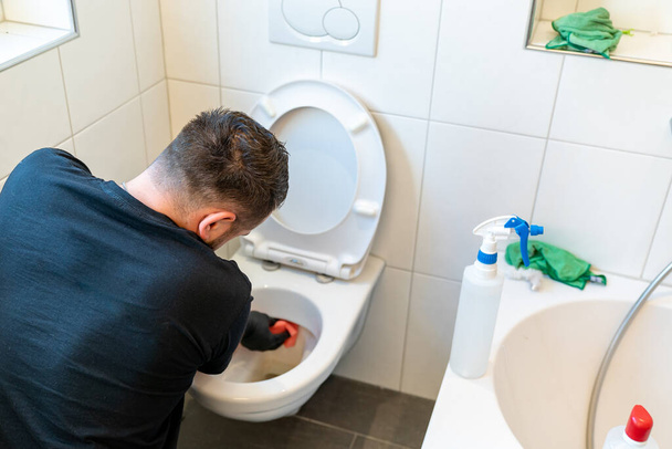 Профессиональный уборщик очень тщательно чистит керамический туалет в современной ванной комнате
 - Фото, изображение