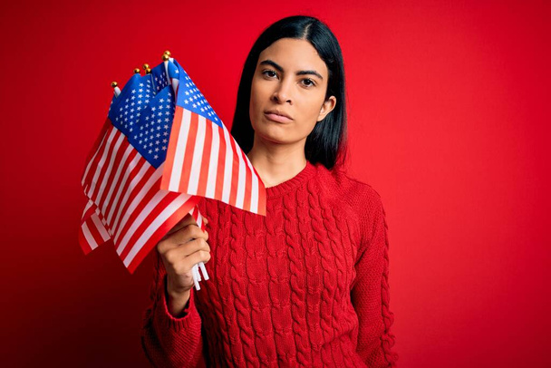 Jovem bela mulher hispânica segurando Estados Unidos da bandeira americana no dia da independência com uma expressão confiante no rosto inteligente pensando sério
 - Foto, Imagem