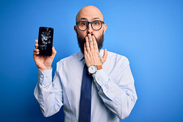 Handsome bald business man with beard holding broken smartphone showing cracked screen cover mouth with hand shocked with shame for mistake, expression of fear, scared in silence, secret concept - Foto, afbeelding