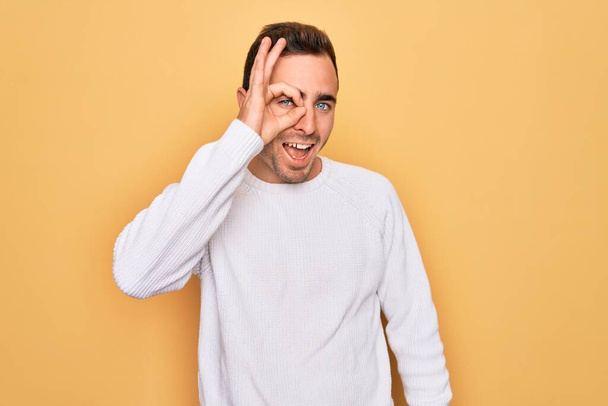 Young handsome man with blue eyes wearing casual sweater standing over yellow background doing ok gesture with hand smiling, eye looking through fingers with happy face. - Fotó, kép