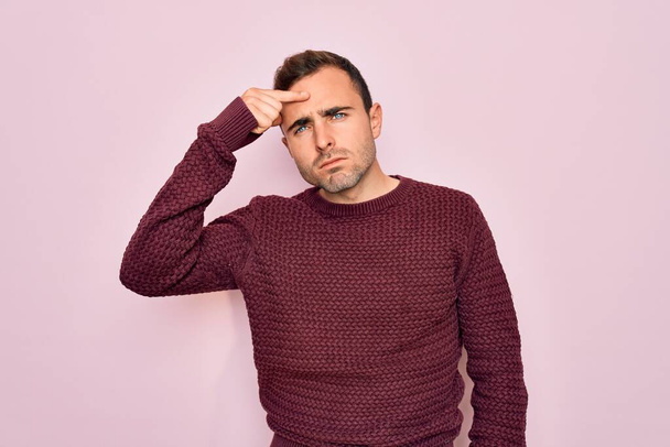 Young handsome man with blue eyes wearing casual sweater standing over pink background pointing unhappy to pimple on forehead, ugly infection of blackhead. Acne and skin problem - Photo, Image