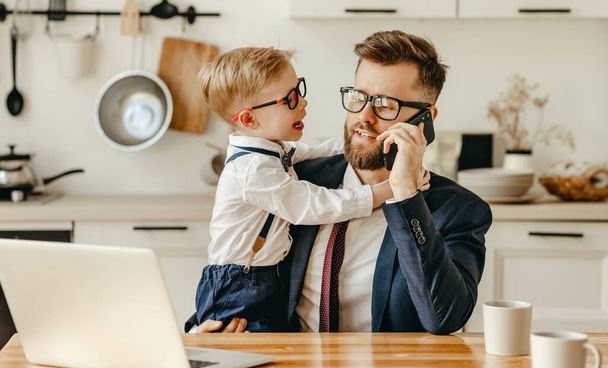 Modern male entrepreneur in formal outfit sitting at table with laptop and speaking on mobile phone while playful son asking for attention during remote work from hom - Photo, Image