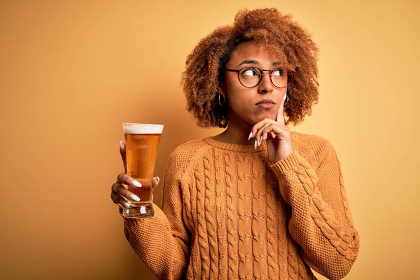 Νεαρή όμορφη Αφρο-Αμερικανίδα με σγουρά μαλλιά πίνοντας ένα ποτήρι μπύρα σοβαρό πρόσωπο σκεπτόμενη την ερώτηση, πολύ μπερδεμένη ιδέα - Φωτογραφία, εικόνα