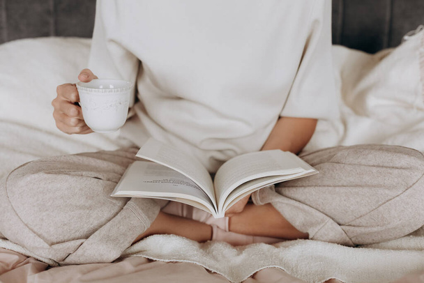 girl in pajamas reads a book and holds a cup of tea inside bedroom - Zdjęcie, obraz