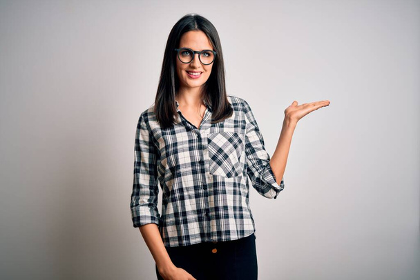 Young brunette woman with blue eyes wearing casual shirt and glasses over white background smiling cheerful presenting and pointing with palm of hand looking at the camera. - 写真・画像