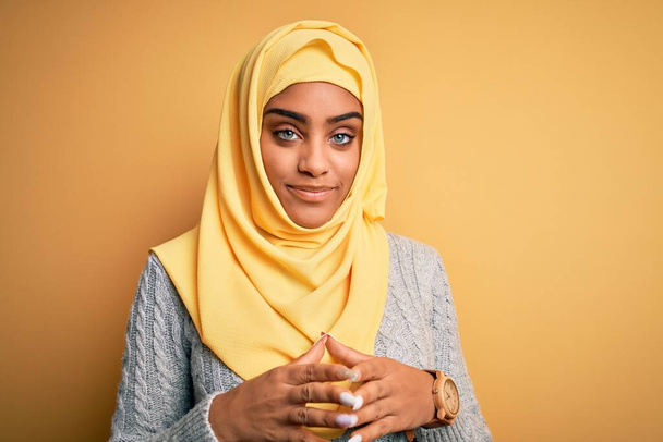 孤立した黄色の背景の上にイスラム教徒のヒジャーブを身に着けている若い美しいアフリカ系アメリカ人の少女手を一緒にし、指はリラックスして陽気に笑顔を渡った。成功と楽観的 - 写真・画像