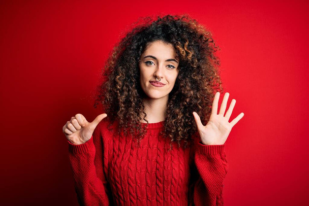 Young beautiful woman with curly hair and piercing wearing casual red sweater showing and pointing up with fingers number six while smiling confident and happy. - Photo, Image