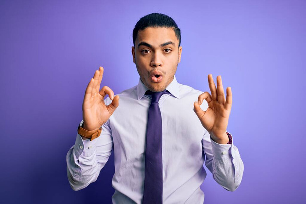Jeune homme d'affaires brésilien portant une cravate élégante debout sur fond violet isolé regardant surpris et choqué faisant ok symbole d'approbation avec les doigts. Expression folle
 - Photo, image