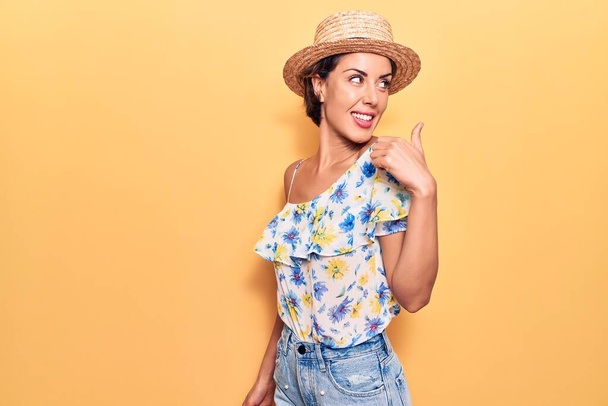 Young beautiful woman wearing summer hat pointing thumb up to the side smiling happy with open mouth  - Foto, imagen