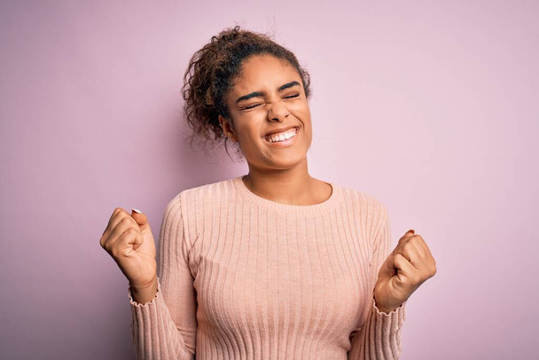 Giovane bella ragazza afroamericana indossa maglione casual in piedi su sfondo rosa molto felice ed eccitato facendo gesto vincitore con le braccia alzate, sorridente e urlando per il successo. Concetto di celebrazione
. - Foto, immagini