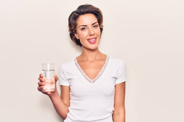 Νεαρή όμορφη γυναίκα πίνοντας ποτήρι νερό φαίνεται θετική και ευτυχισμένη στέκεται και χαμογελά με αυτοπεποίθηση χαμόγελο δείχνει τα δόντια  - Φωτογραφία, εικόνα