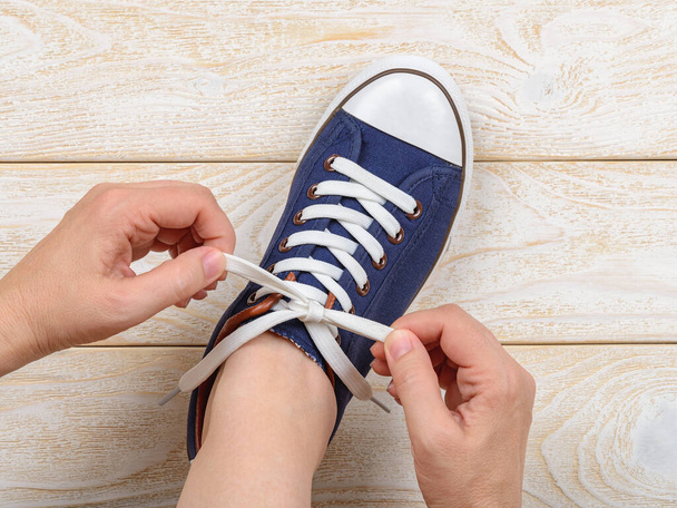 Caucasian woman hands tie white elastic laces on classic blue  gumshoes or sneakers indoor. Comfortable shoes for fitness and sport. Shoes with lacing. Top view. - Foto, Imagen