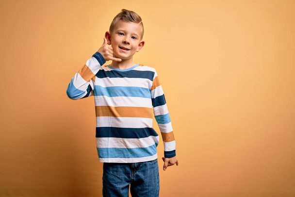 Jeune enfant caucasien aux yeux bleus portant une chemise rayée colorée sur fond jaune souriant faisant geste téléphonique avec la main et les doigts comme parler au téléphone. Communication des concepts
. - Photo, image