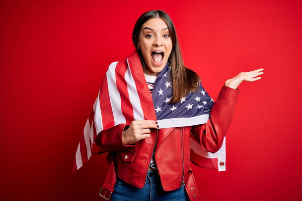 Junge schöne brünette patriotische Frau trägt Flagge der Vereinigten Staaten für den Unabhängigkeitstag sehr glücklich und aufgeregt, Siegerausdruck feiert Sieg schreiend mit großem Lächeln und erhobenen Händen - Foto, Bild
