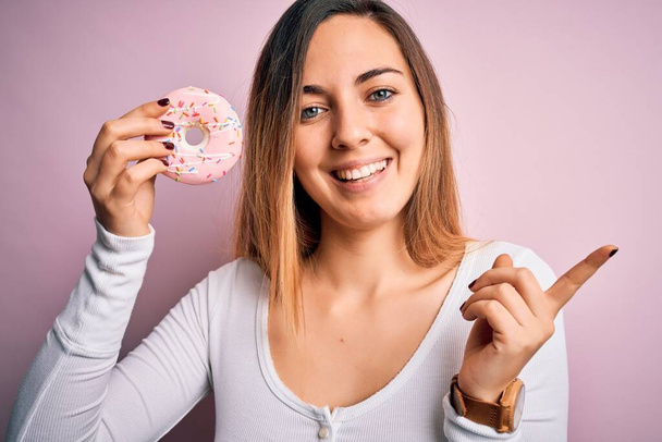 jong mooi blond vrouw met blauwe ogen houden roze donut over geïsoleerde achtergrond zeer gelukkig wijzend met hand en vinger naar de zijkant - Foto, afbeelding