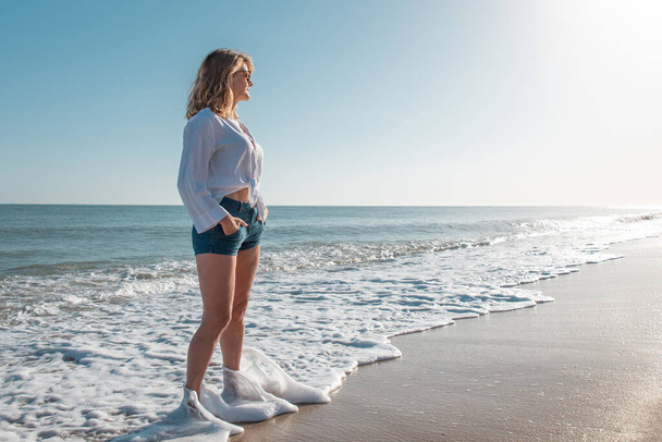 Mulher olhando para a paisagem do mar na praia em uma camisa branca
 - Foto, Imagem
