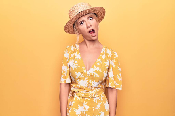 Belle femme blonde en vacances portant un chapeau et une robe d'été sur fond jaune Dans un visage choqué, sceptique et sarcastique, surpris avec la bouche ouverte
 - Photo, image
