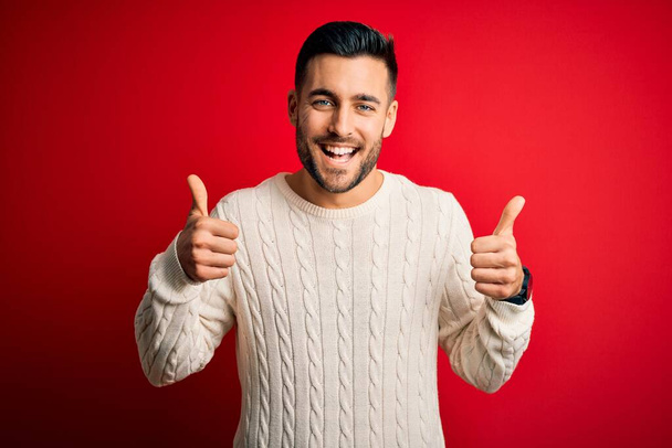 Jonge knappe man draagt casual witte trui staan over geïsoleerde rode achtergrond succes teken doen positieve gebaar met de hand, duimen omhoog glimlachend en gelukkig. Vrolijke uitdrukking en winnaar gebaar. - Foto, afbeelding