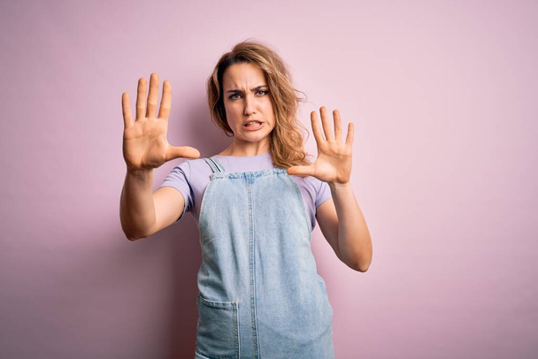 Young beautiful blonde woman wearing casual denim overalls standing over pink background afraid and terrified with fear expression stop gesture with hands, shouting in shock. Panic concept. - Foto, immagini