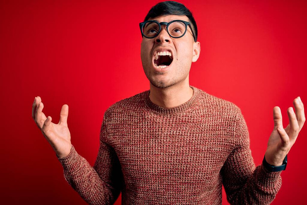 Homem hispânico bonito jovem usando óculos nerd sobre fundo vermelho louco e louco gritando e gritando com expressão agressiva e braços levantados. Conceito de frustração
. - Foto, Imagem