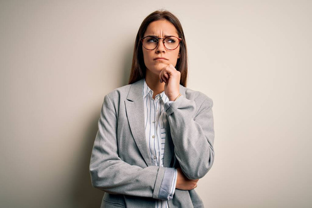 Young beautiful brunette businesswoman wearing jacket and glasses over white background with hand on chin thinking about question, pensive expression. Smiling with thoughtful face. Doubt concept. - Photo, Image