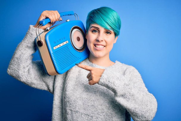 Jonge vrouw met blauwe mode haar luisteren naar muziek houden vintage draagbare radio zeer gelukkig wijzend met hand en vinger - Foto, afbeelding