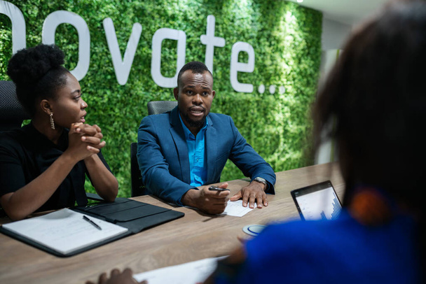 Jonge Afrikaanse zakenman bespreekt werk met een groep collega 's tijdens een bijeenkomst rond een tafel in een kantoordirectiekamer - Foto, afbeelding