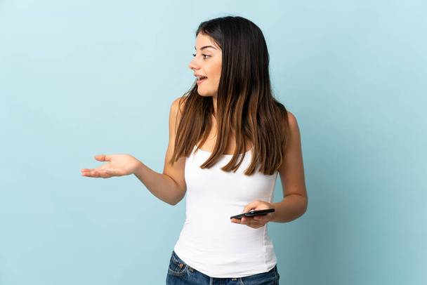 Молода біла жінка використовує мобільний телефон ізольовано на синьому фоні з несподіваним виразом, дивлячись збоку
 - Фото, зображення