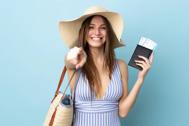 Joven mujer caucásica en vacaciones de verano sosteniendo pasaporte aislado sobre fondo azul señala el dedo en usted con una expresión de confianza
 - Foto, imagen