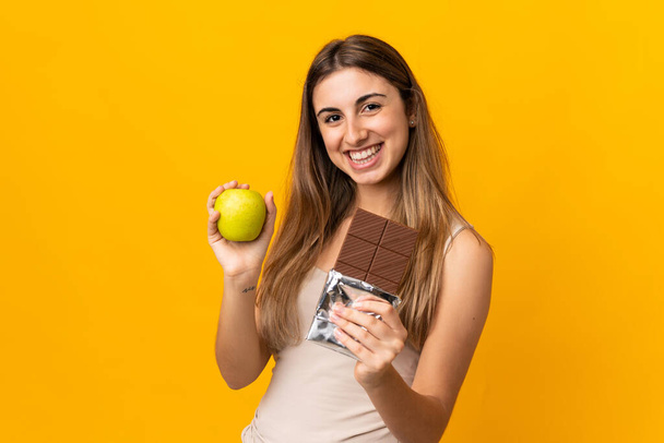 Νεαρή γυναίκα πάνω από απομονωμένο κίτρινο φόντο λαμβάνοντας ένα δισκίο σοκολάτας στο ένα χέρι και ένα μήλο στο άλλο - Φωτογραφία, εικόνα