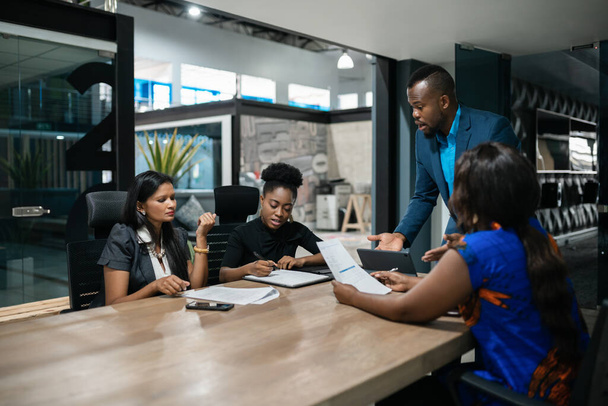 Jonge Afrikaanse zakenman in gesprek met een divers team van collega 's tijdens een bijeenkomst rond een tafel in een kantoor bestuurskamer - Foto, afbeelding