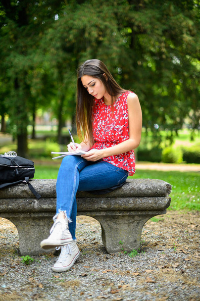 Красивая студентка колледжа читает книгу на скамейке в парке - Фото, изображение