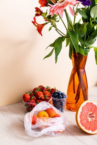 floral and fruit composition with bouquet in vase, berries, grapefruit and apricots on wooden surface on beige background - Photo, Image