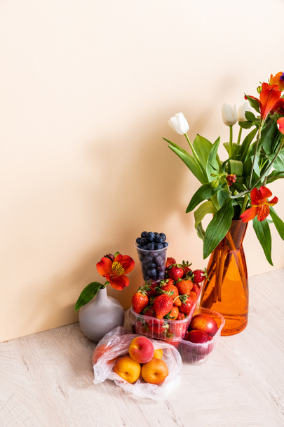 floral and fruit composition with bouquet in vase and summer fruits on wooden surface on beige background - Photo, Image