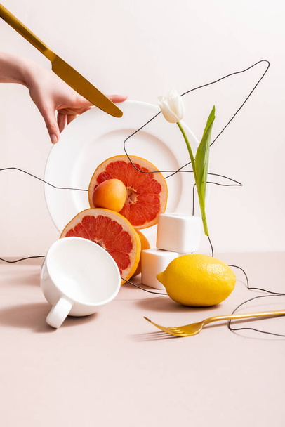 composição floral e frutífera com tulipa em fio e frutas cítricas perto de cubos, xícara, garfo, faca e mão feminina em placa isolada em bege
 - Foto, Imagem