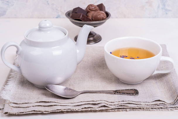Taza de té verde con pétalos de flores en una servilleta de lino y caramelos en un jarrón de metal vintage sobre una mesa de madera blanca, primer plano
 - Foto, Imagen